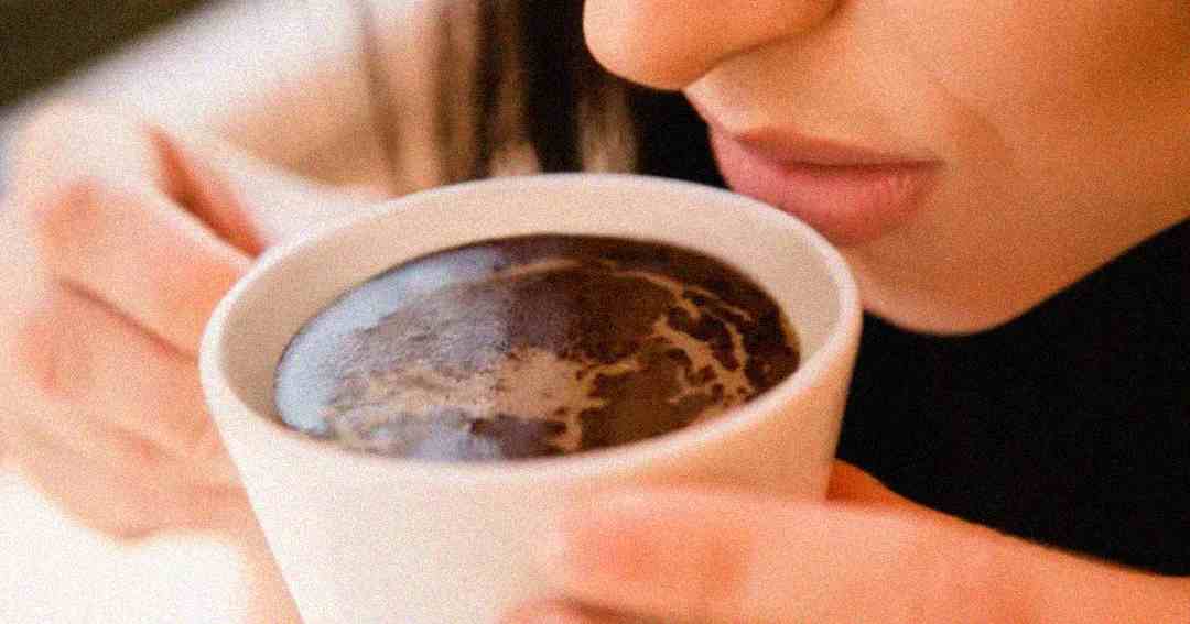 A close-up of a person holding a white cup of dark coffee near their lips, about to take a sip. The steam and froth are visible on the surface of the coffee.