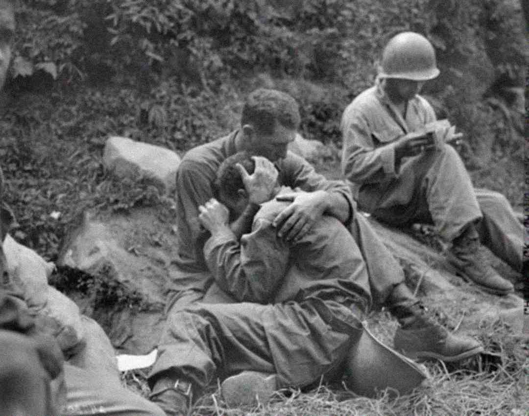 Two soldiers sit on the ground; one cradles and comforts the other, who is visibly distressed. Another soldier sits nearby, looking down at an object in his hands, wearing a helmet. The mood is somber and emotional.