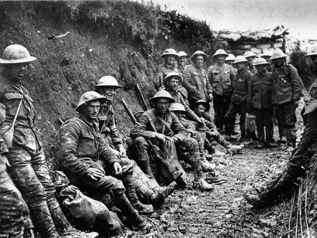 A group of World War I soldiers in uniforms and helmets sit and stand along a muddy trench, some holding rifles, appearing tired and somber. The background shows rough earth walls and overcast sky.