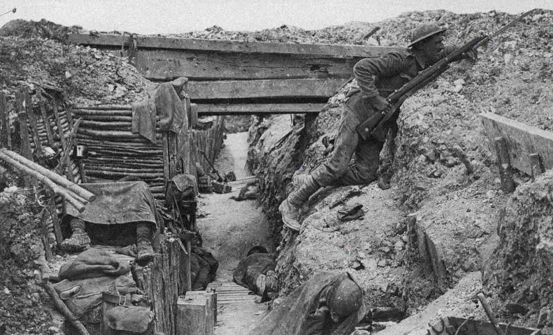 A black-and-white photo of WWI soldiers in a muddy trench. One soldier with a rifle climbs over the trench's edge, while others lie low or take cover among scattered gear and rough wooden supports.