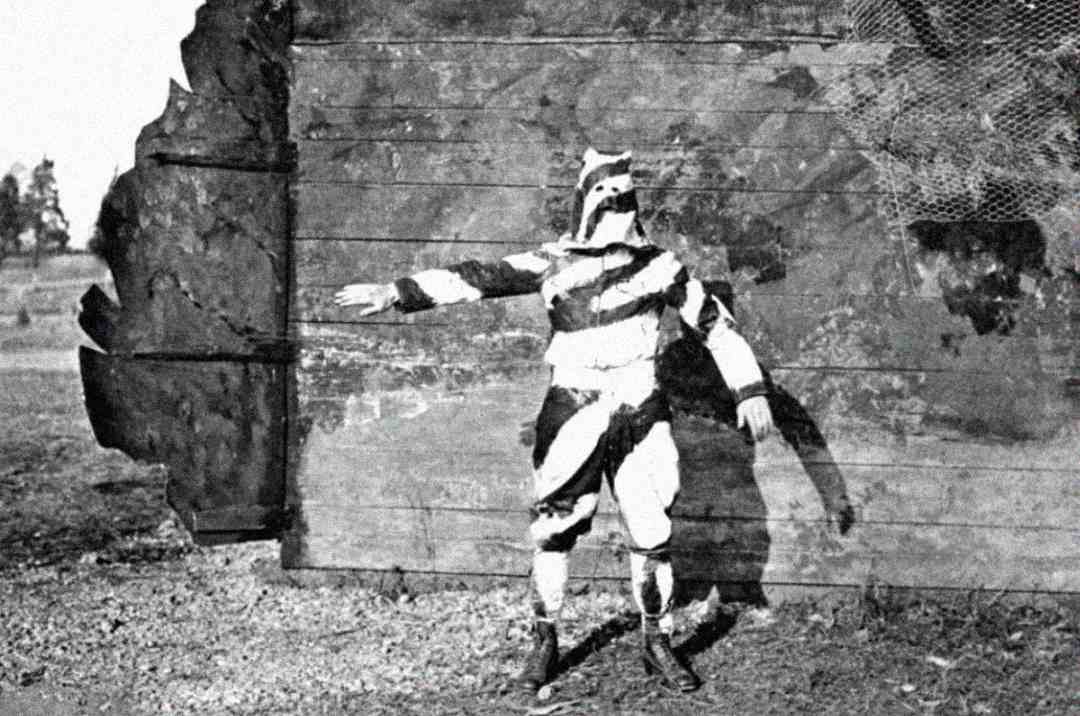 A person dressed in a black-and-white striped costume stands with one arm raised in front of a large wooden structure outdoors. The costume covers their entire body, including their face.