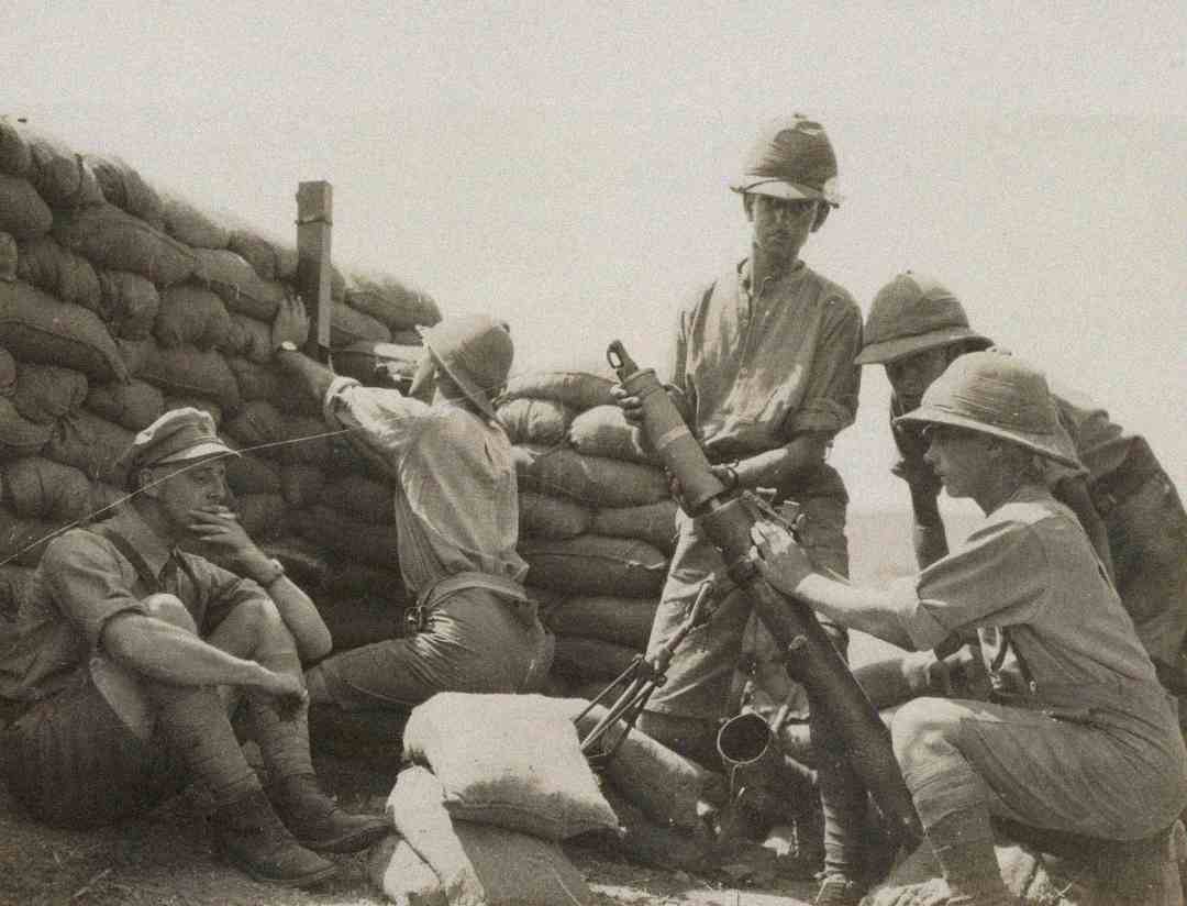 Five soldiers in pith helmets operate a mortar behind a sandbag barrier, likely during a historical military conflict. Some are adjusting the mortar while others look on or prepare ammunition.