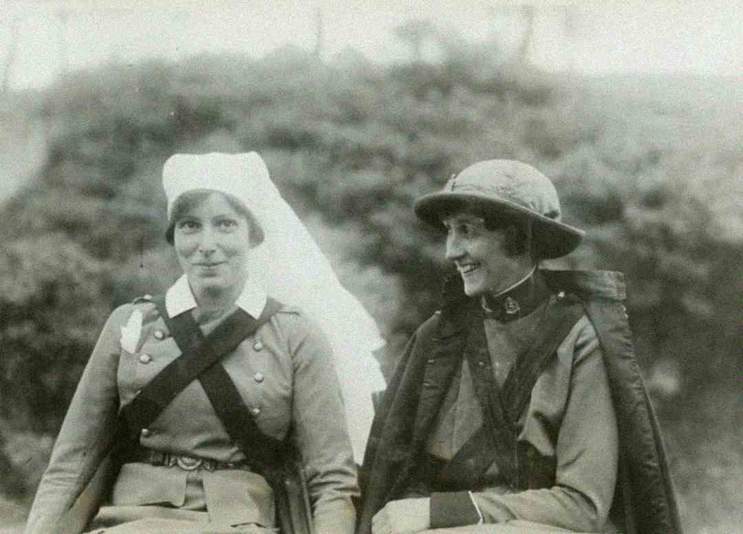Two women in World War I-era uniforms sit side by side outdoors, one wearing a nurse's outfit with a white veil, and the other in military attire with a helmet, both smiling.