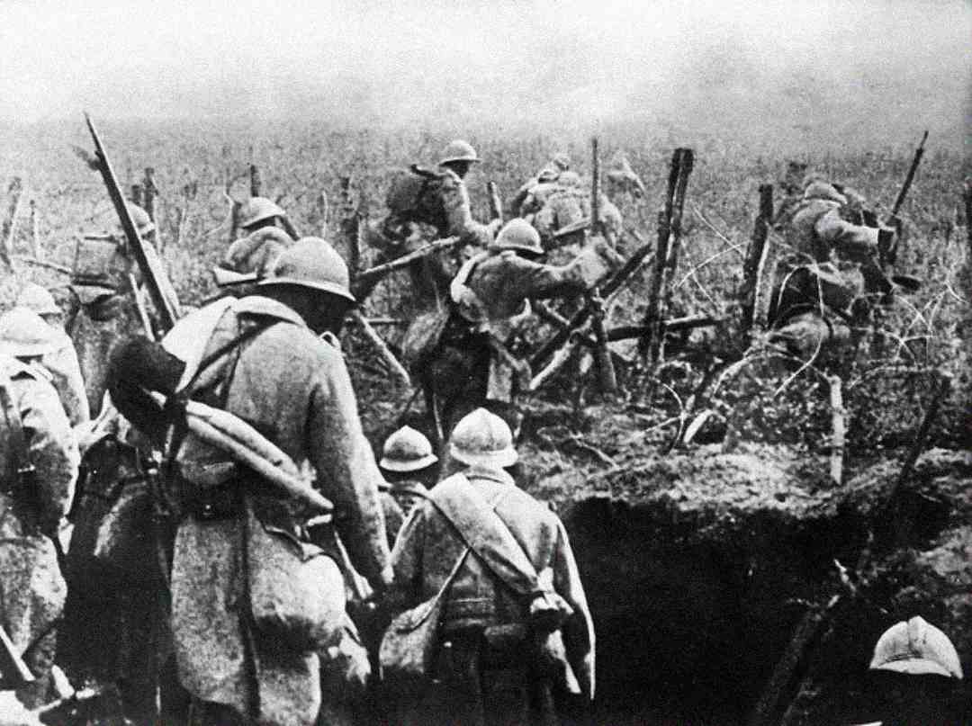 Black and white photo of soldiers in World War I uniforms and helmets climbing out of a trench and advancing through barbed wire on a battlefield, holding rifles and equipment.