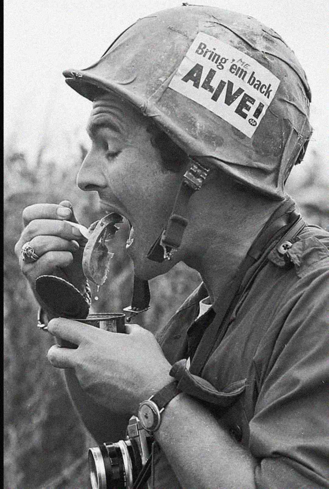 A soldier wearing a helmet with a "Bring 'em back ALIVE!" sign eats from a can with a spoon, holding a camera around his neck and a ring on his finger. The photo is in black and white.