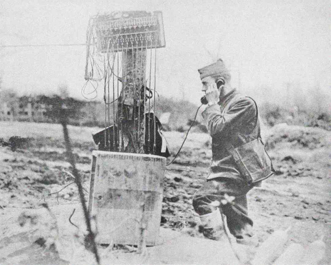A soldier kneels and speaks into a field telephone connected to a makeshift communication post with exposed wires in a barren, war-torn landscape.