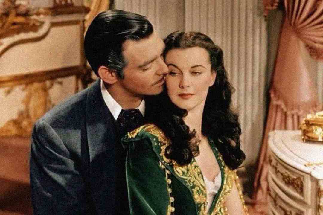 A man in a suit gently leans toward a woman in a green dress with gold details. She looks away with a calm expression as they sit closely together in an elegant, vintage room with ornate decor.