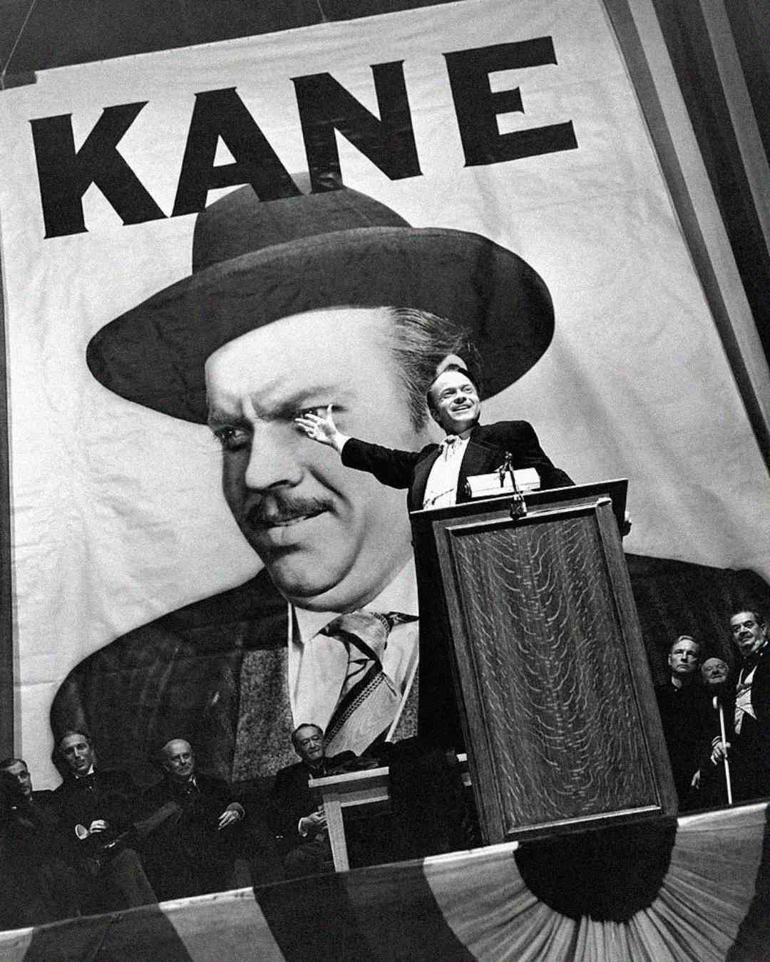 A man stands at a podium, gesturing passionately, in front of a giant poster of himself labeled "KANE." Several people are seated behind him on stage. The scene suggests a political rally or speech.
