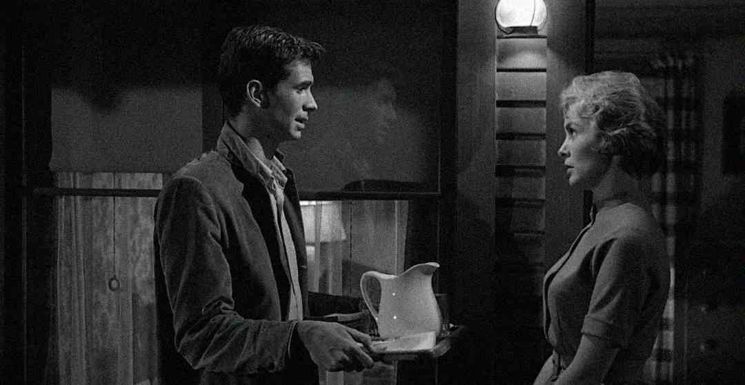 A man holding a tray with a pitcher speaks to a woman outside a building at night, illuminated by a single porch light. The scene is in black and white, creating a dramatic atmosphere.