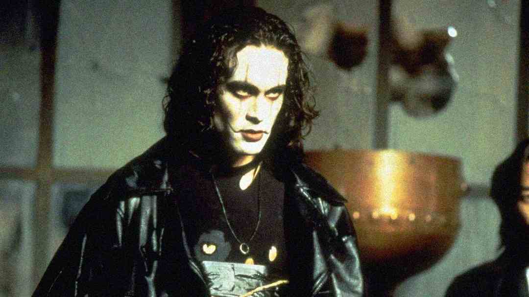 A person with long dark hair and white face paint wears a black leather coat and dark clothing, standing indoors with a somber expression. The background is dimly lit and slightly out of focus.