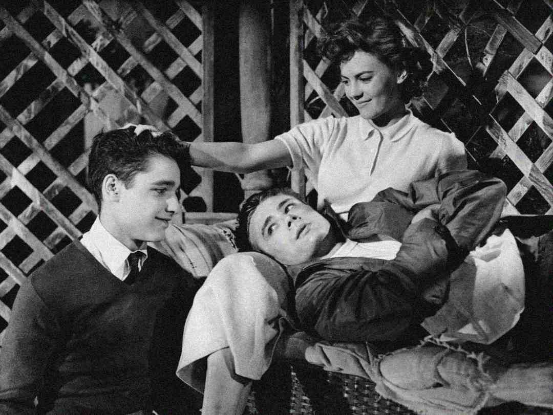 Three young people sit close together. A woman gently touches the hair of a boy lying across a bench, while another boy sits nearby, looking at them. The scene appears to be from an old black-and-white film.