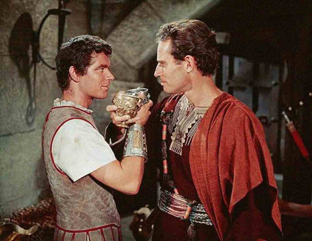 Two men dressed in ancient Roman-style costumes, one holding a decorative cup up to the other’s lips, stand closely together in a dimly lit room with stone walls and armor in the background.