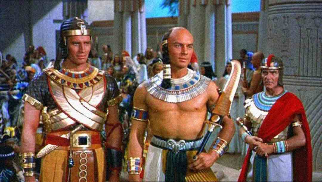Three men in elaborate ancient Egyptian costumes stand indoors among others; two wear gold and jeweled armor, one shirtless with a wide collar, and all are surrounded by Egyptian-style decor and people in colorful attire.