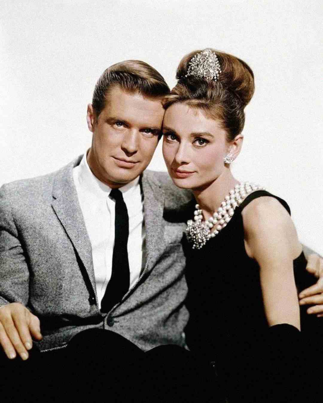 A man in a gray suit and a woman in a black dress with a pearl necklace and jeweled hairpiece sit closely together, facing the camera against a plain background.