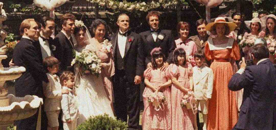 A large group of people poses for a formal wedding photo outdoors. The bride and groom stand in the center, surrounded by men in tuxedos, women in dresses, and several flower girls in pink dresses.