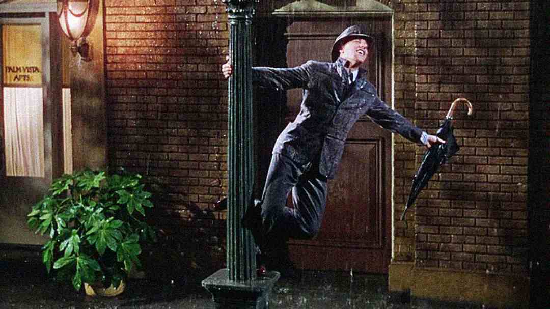 A man in a suit and hat joyfully swings from a lamppost with one arm, holding an umbrella in the other hand, as rain pours down on a city street at night.
