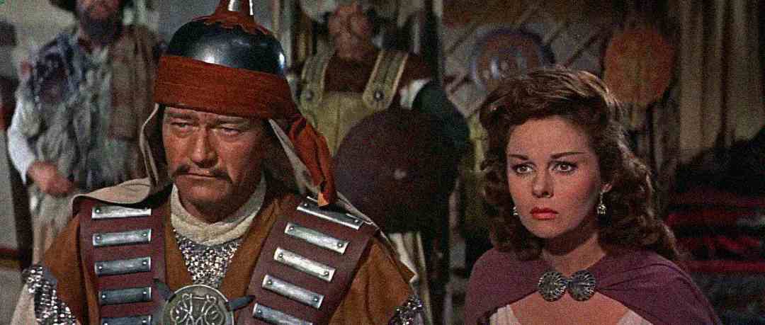 A man in armor and a helmet with a stern expression stands next to a woman with brown hair in a purple cloak, both appearing serious, with other people blurred in the background.
