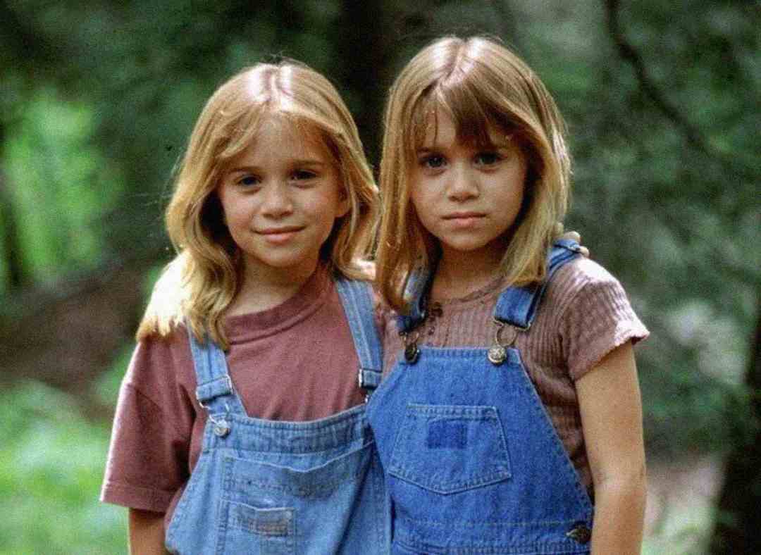 Two young girls with long blonde hair wearing denim overalls and t-shirts stand side by side outdoors, with one girl's arm around the other, in a green, natural setting.