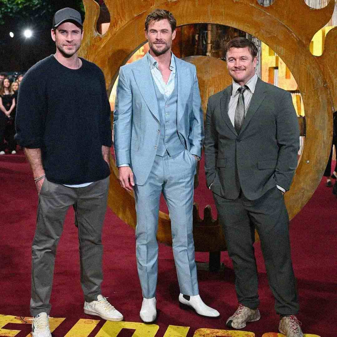 Three men stand together on a red carpet in front of a large circular backdrop. The man in the center wears a light blue suit; the other two wear dark suits. All three are smiling and posing for the photo.