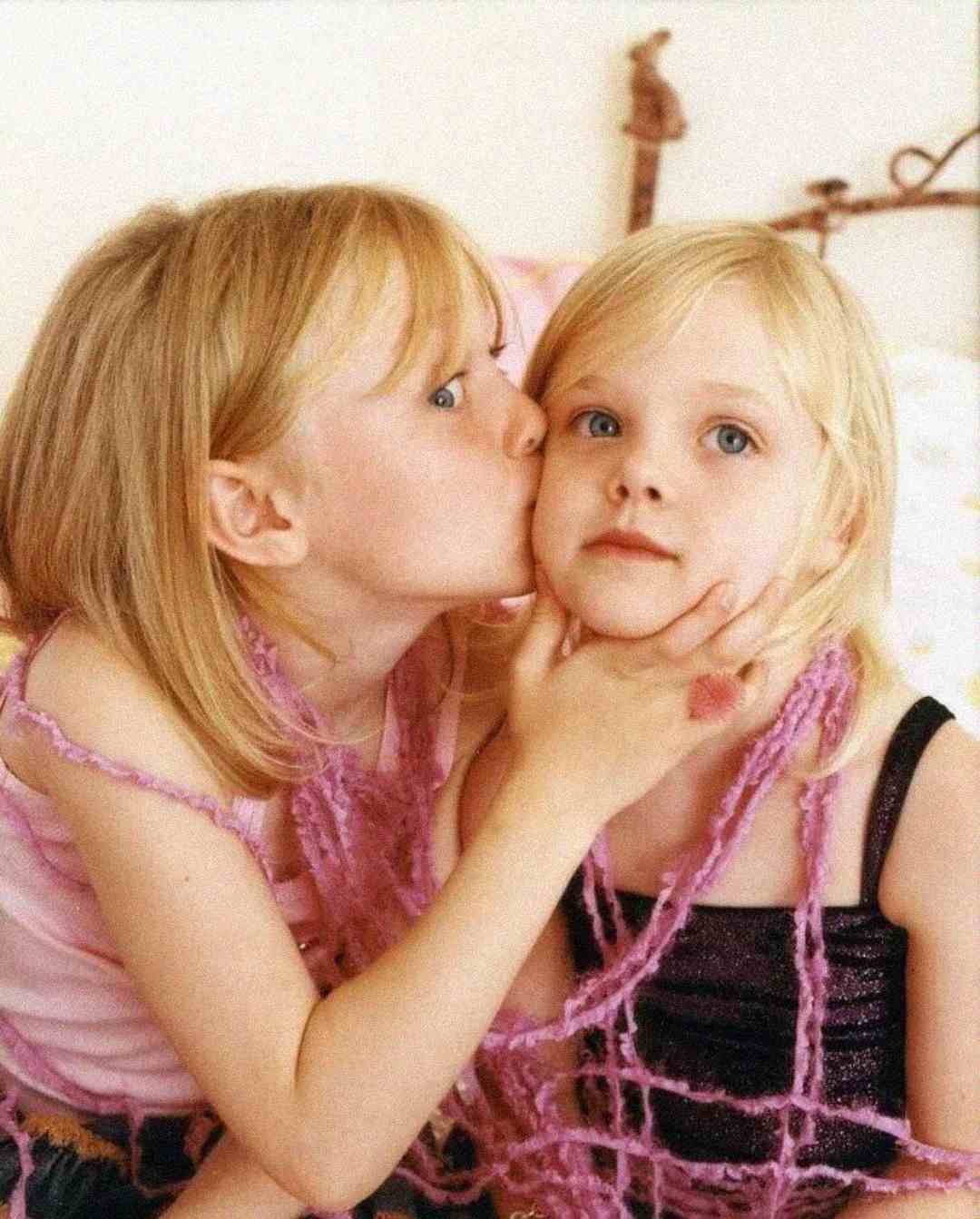 Two young blonde girls sit close together. One girl kisses the other on the cheek and gently holds her face, while the second girl looks forward with a neutral expression. Both wear pink and purple outfits.