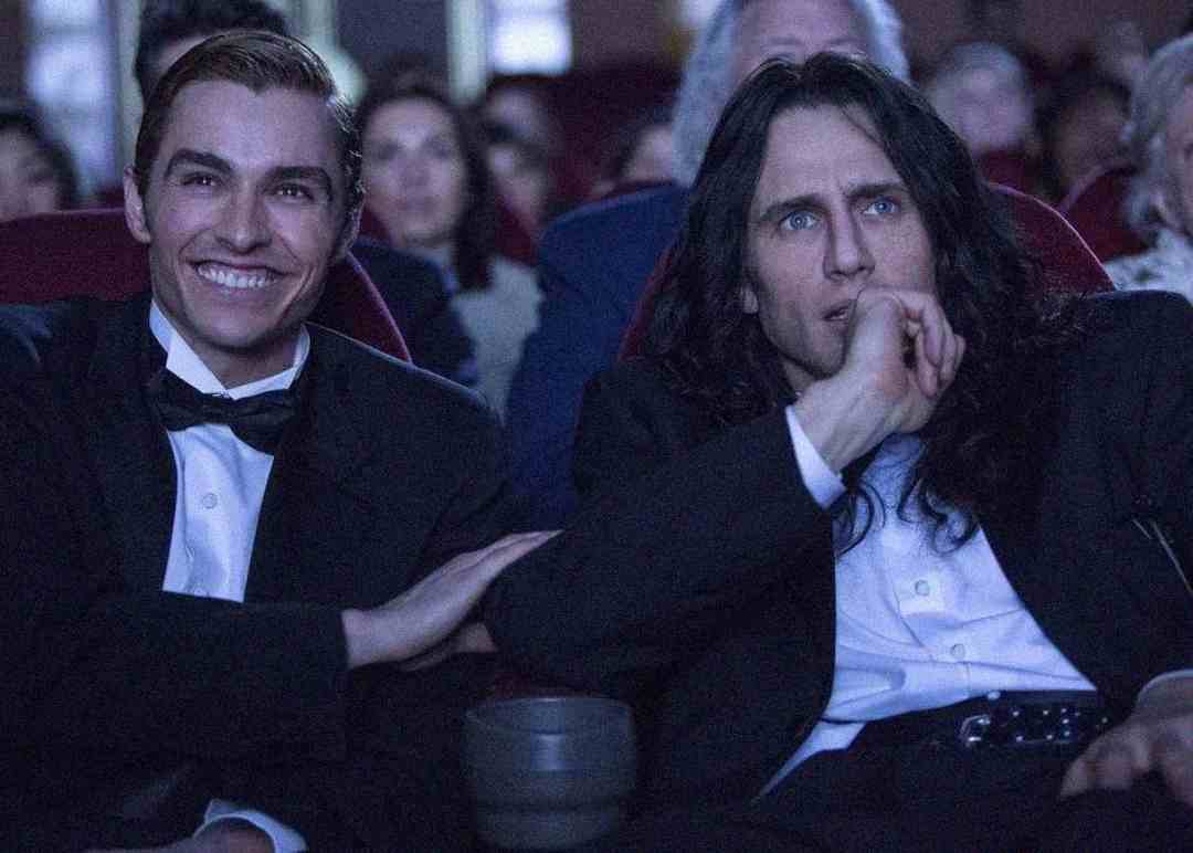 Two men in tuxedos sit in a theater. One smiles and touches the other's arm, while the other looks ahead with a thoughtful, tense expression. Both are surrounded by other seated people in dim lighting.