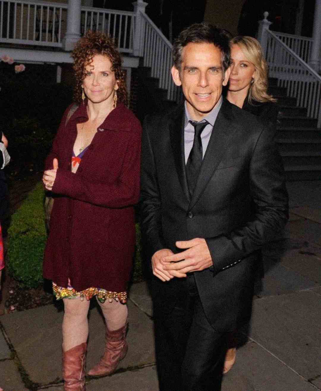 A man in a dark suit stands outside on a stone path, smiling at the camera, with two women behind him; one woman wears a maroon cardigan and patterned dress, the other wears a black coat. White porch railings are in the background.
