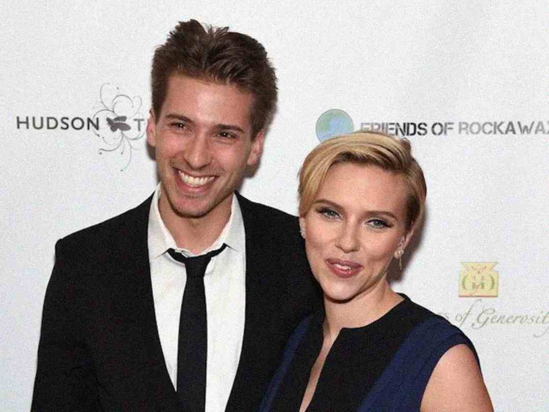 A man in a black suit and tie stands smiling next to a woman with short blonde hair in a dark dress. They are posing in front of a white backdrop with logos and text.