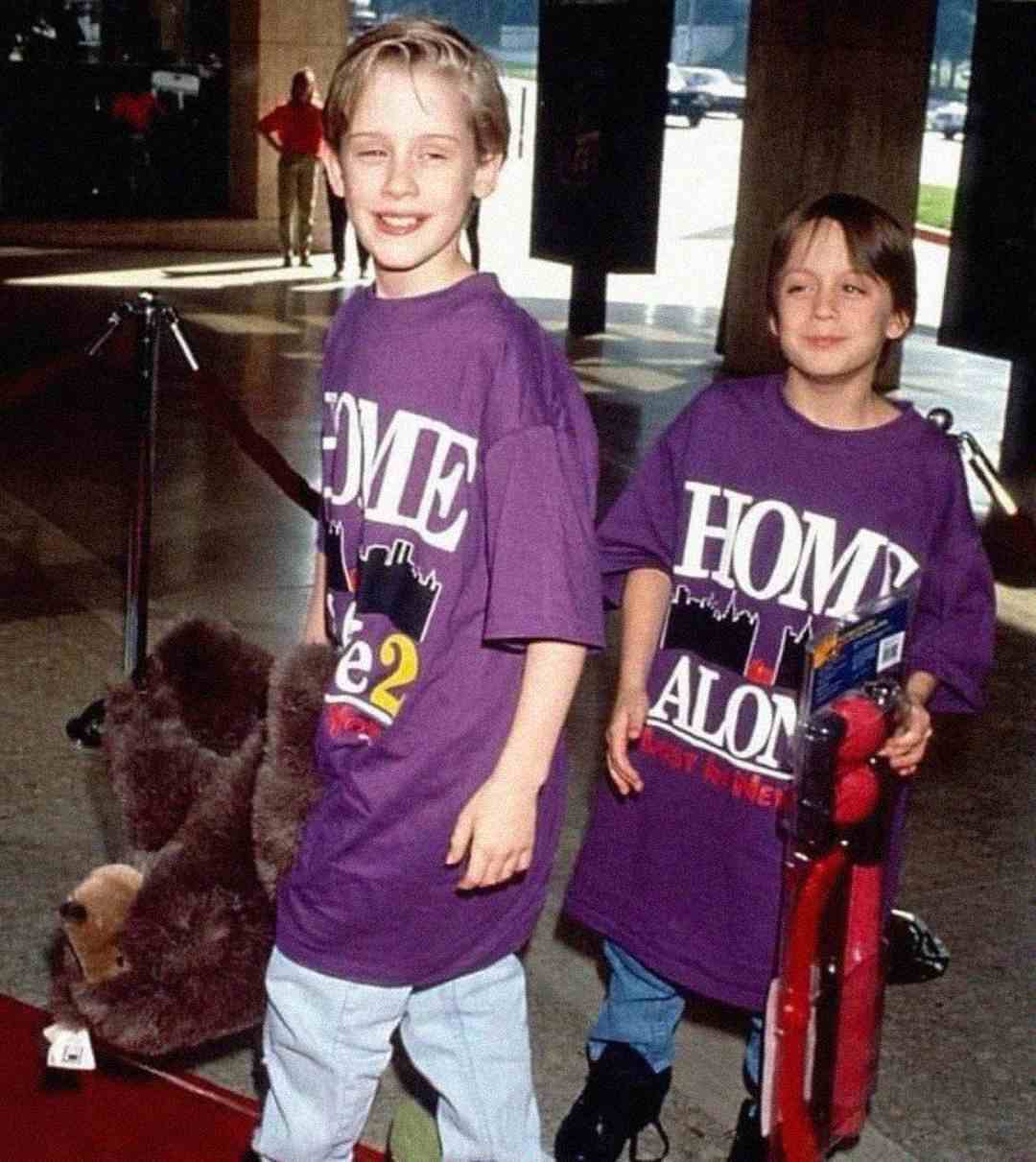 Two smiling children wearing oversized purple "Home Alone 2" T-shirts stand on a red carpet, one holding a large stuffed bear and the other holding a red scooter.
