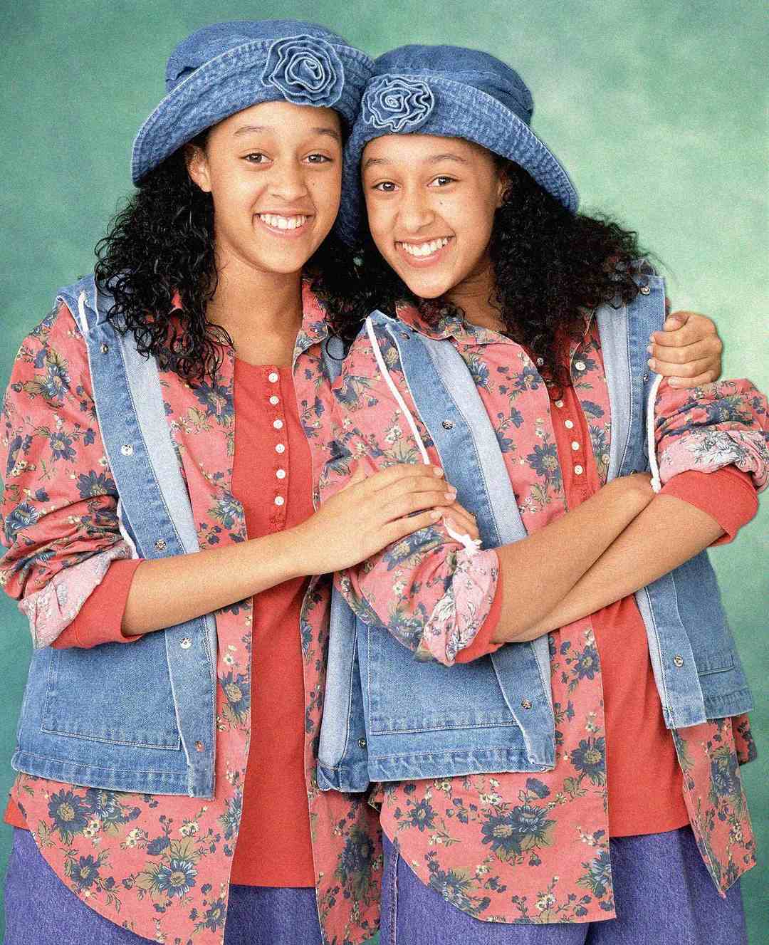 Twin girls with curly hair, wearing matching blue hats, denim vests, and floral shirts, stand close together, smiling and posing in front of a green backdrop.