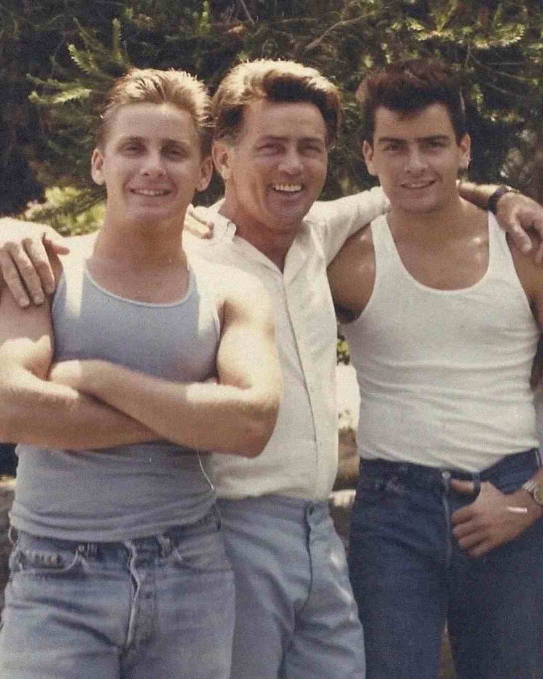 Three men stand closely together outdoors, smiling at the camera. The two younger men wear tank tops and jeans, while the older man in the middle wears a light shirt and pants. Green foliage is visible in the background.