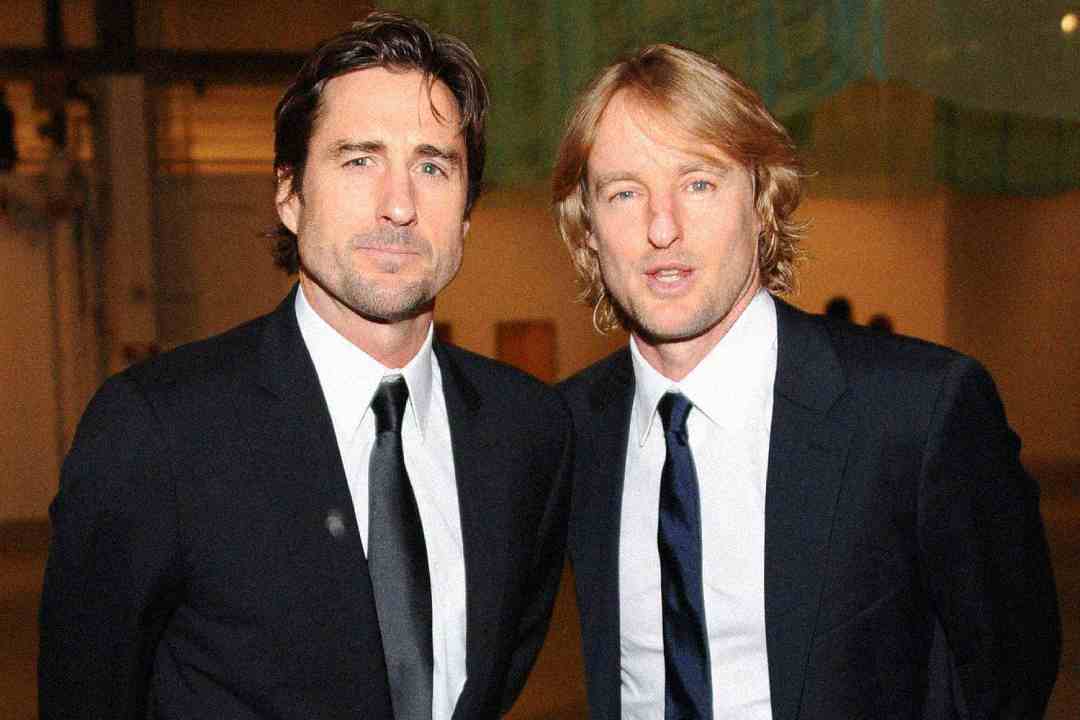 Two men in dark suits and ties stand side by side, facing the camera. They are in an indoor setting with a softly lit background. Both have light facial hair and medium-length hair.