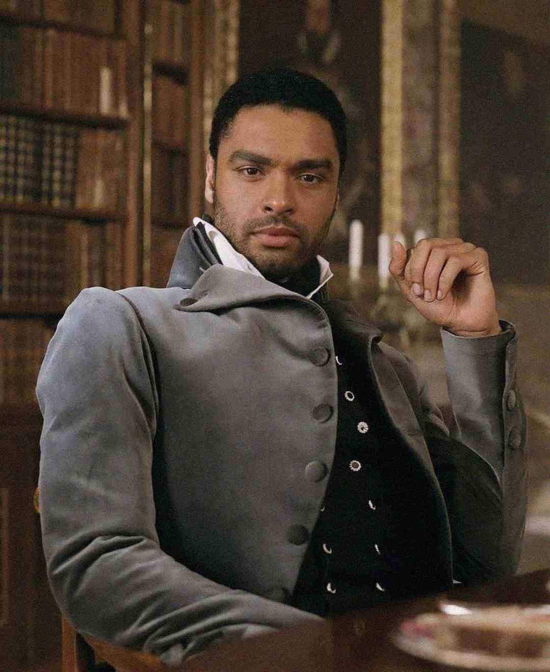 A man in a gray period costume sits at a table in a richly decorated room with bookshelves in the background, looking directly at the camera with a serious expression.
