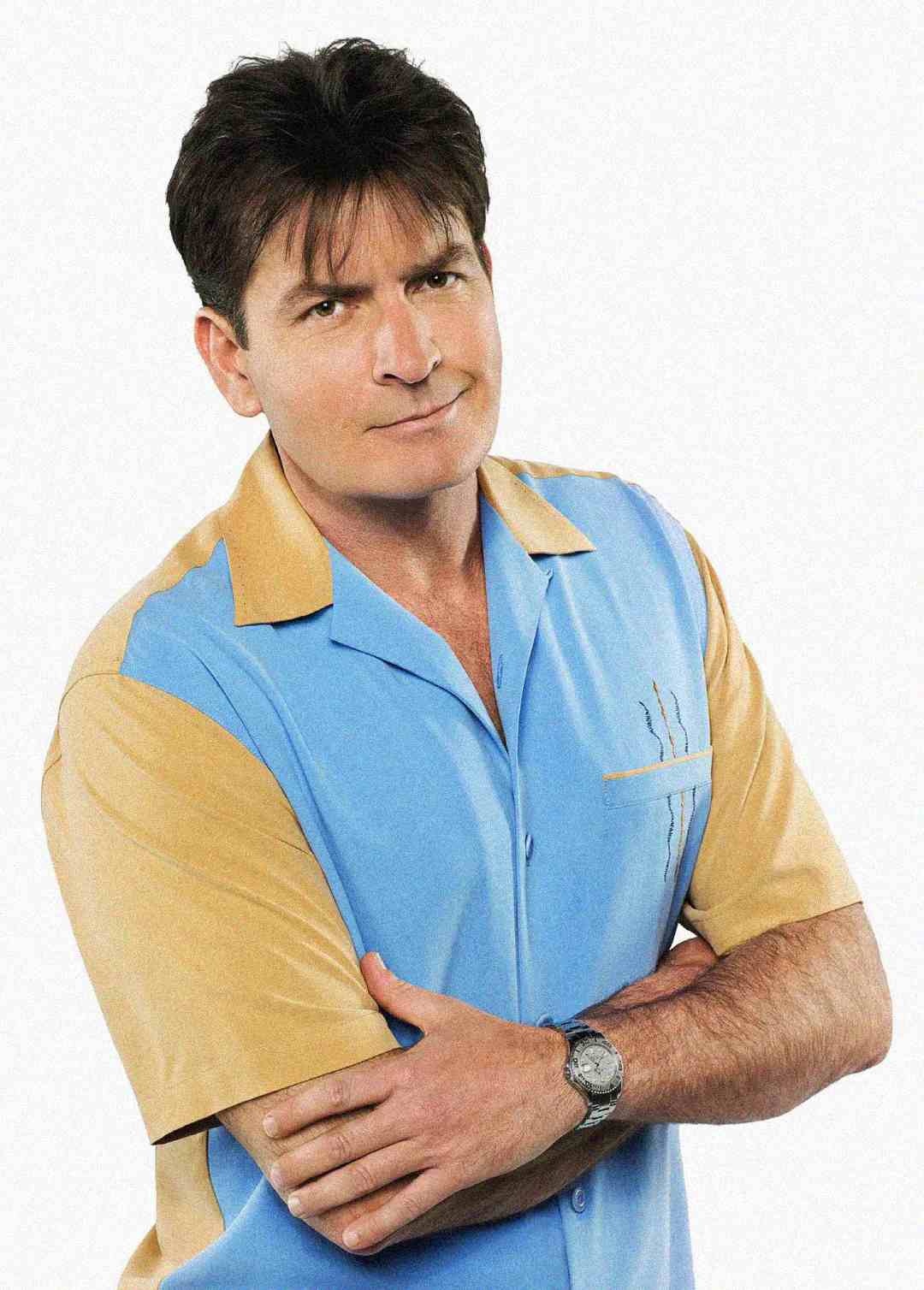 A man with dark hair wearing a blue and tan short-sleeve button-up shirt stands with arms crossed, looking at the camera with a neutral expression.