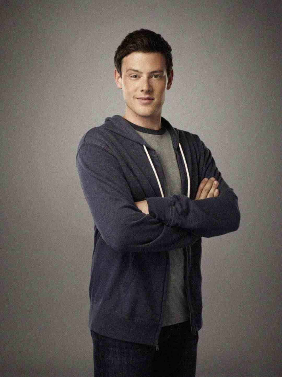 A young man with short brown hair wearing a blue zip-up hoodie over a gray t-shirt stands against a plain gray background with his arms crossed and a slight smile.