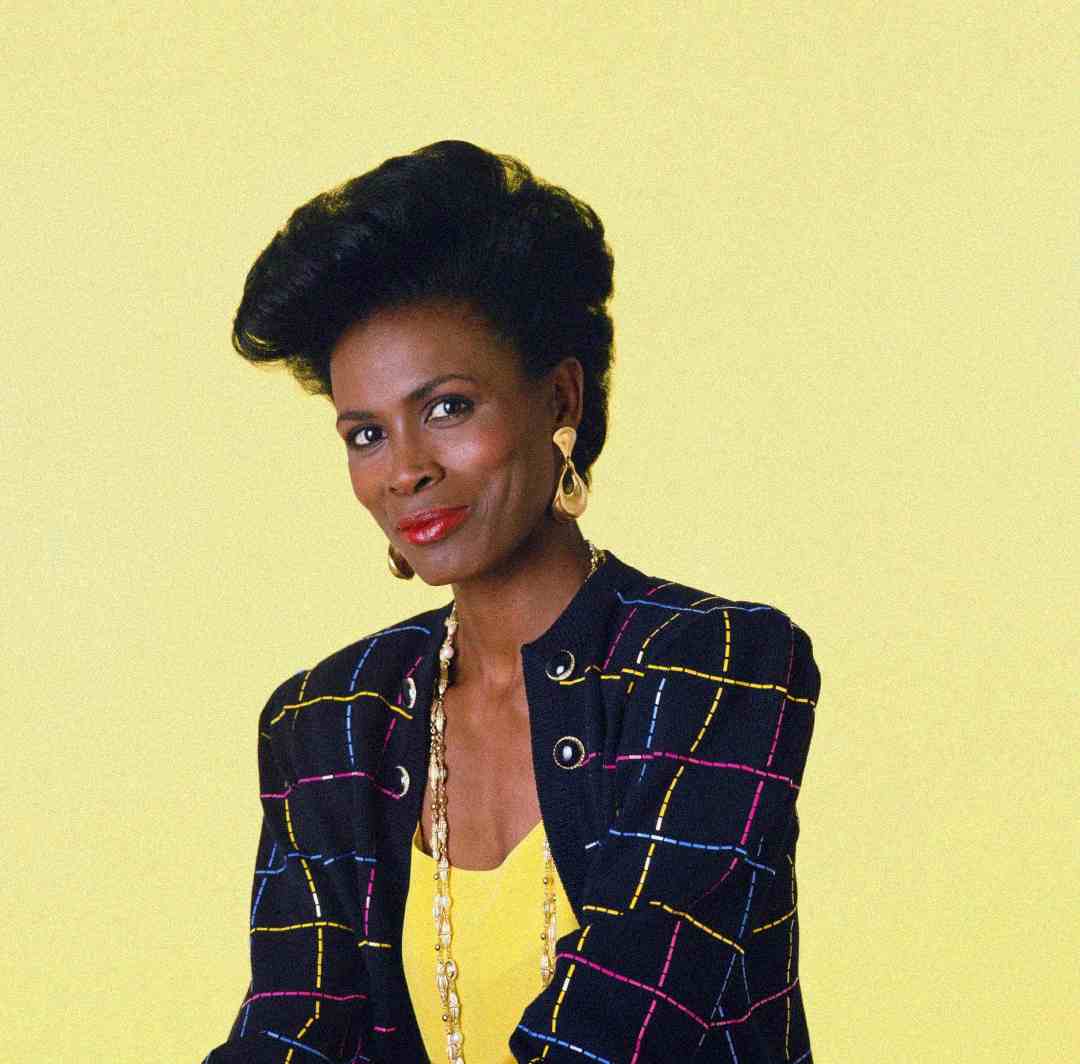 A woman with styled black hair wears a colorful plaid-patterned blazer over a yellow top, gold earrings, and a long necklace, posing against a plain yellow background.