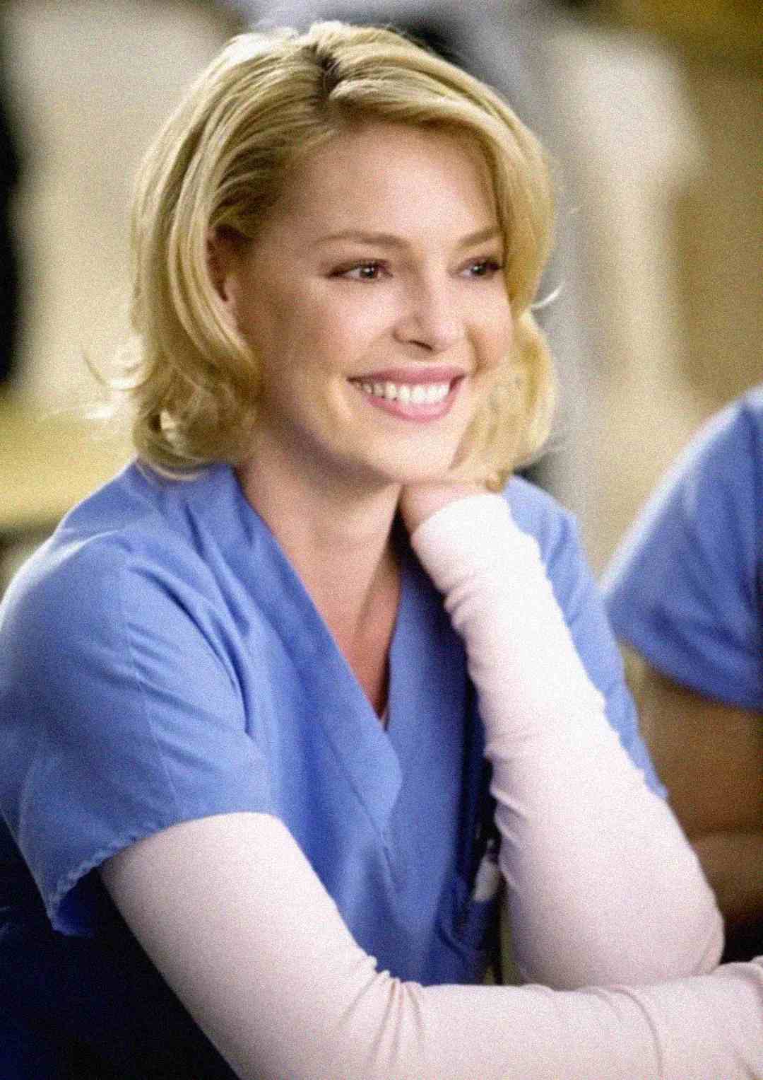 A woman with blonde hair wearing blue medical scrubs and a light pink undershirt is smiling warmly, resting her chin on her hand. The background is softly blurred.