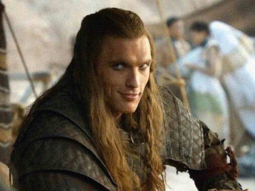 A man with long blond hair and pale skin wears dark, medieval-style armor. He has a sly smile and intense eyes, with a blurred background showing people in light clothing.