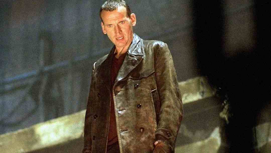 A man with short hair wearing a dark leather jacket stands on a stone staircase, looking slightly downward with a serious expression in a dimly lit setting.