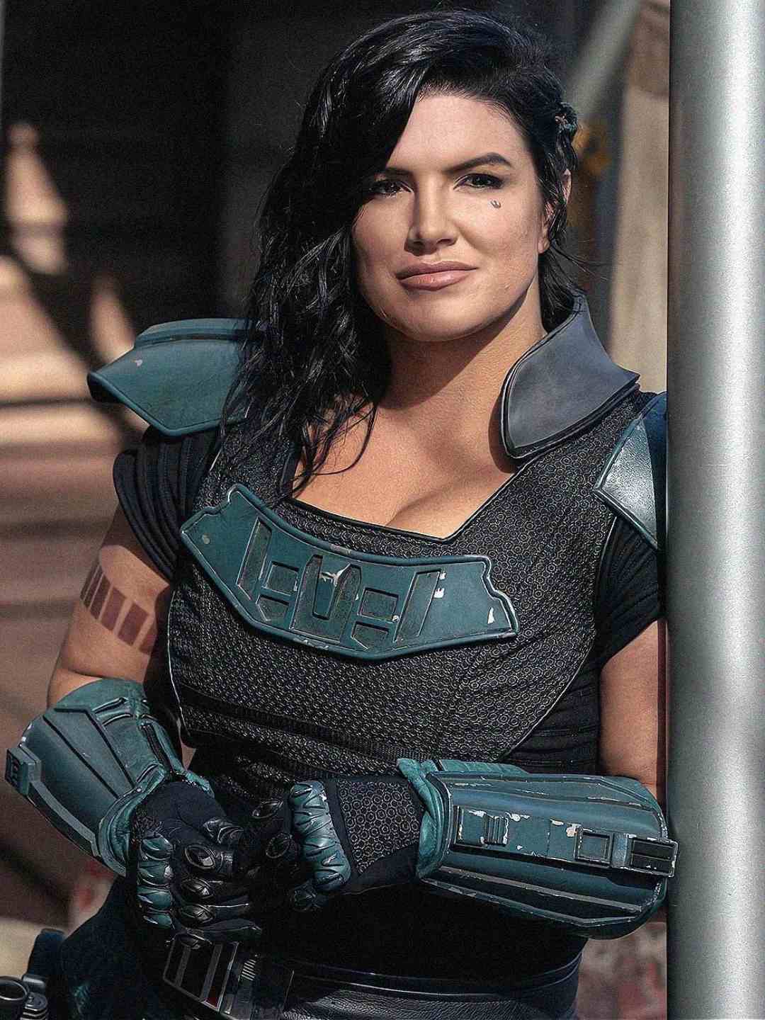 A woman in futuristic black and teal armor with dark wavy hair leans against a metal pole, looking confidently at the camera; she wears gloves and sports a small facial tattoo.