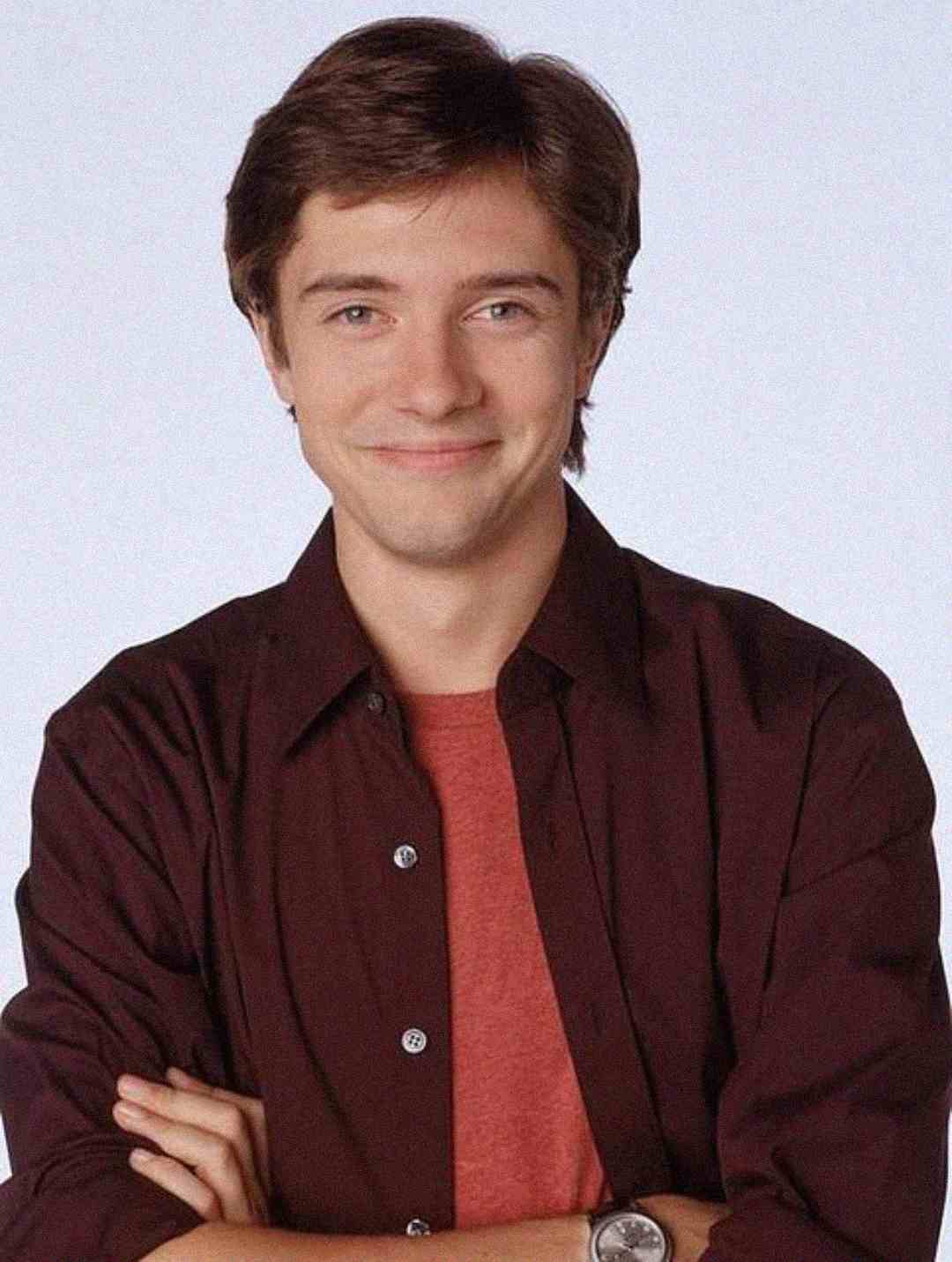 A young man with short brown hair smiles at the camera. He is wearing a maroon button-up shirt over a red t-shirt and has his arms crossed. The background is plain and light-colored.