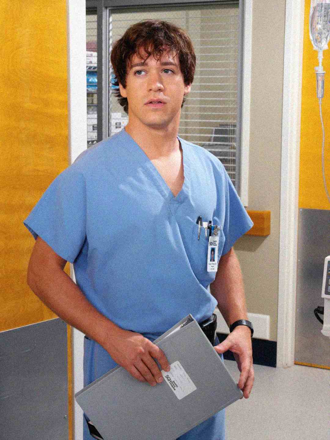 A young male healthcare professional in light blue scrubs stands in a hospital hallway, holding a gray clipboard. Pens and an ID badge are visible in his chest pocket. He looks slightly to the side with a thoughtful expression.