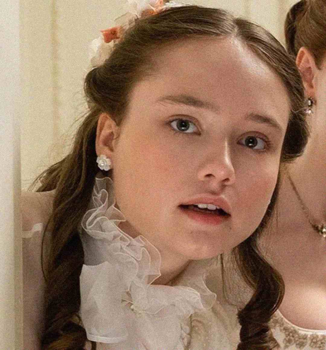 A young woman with fair skin and light brown hair styled in loose curls looks slightly surprised. She wears a white ruffled blouse, pearl earrings, and a floral hair accessory. Another person is partially visible behind her.