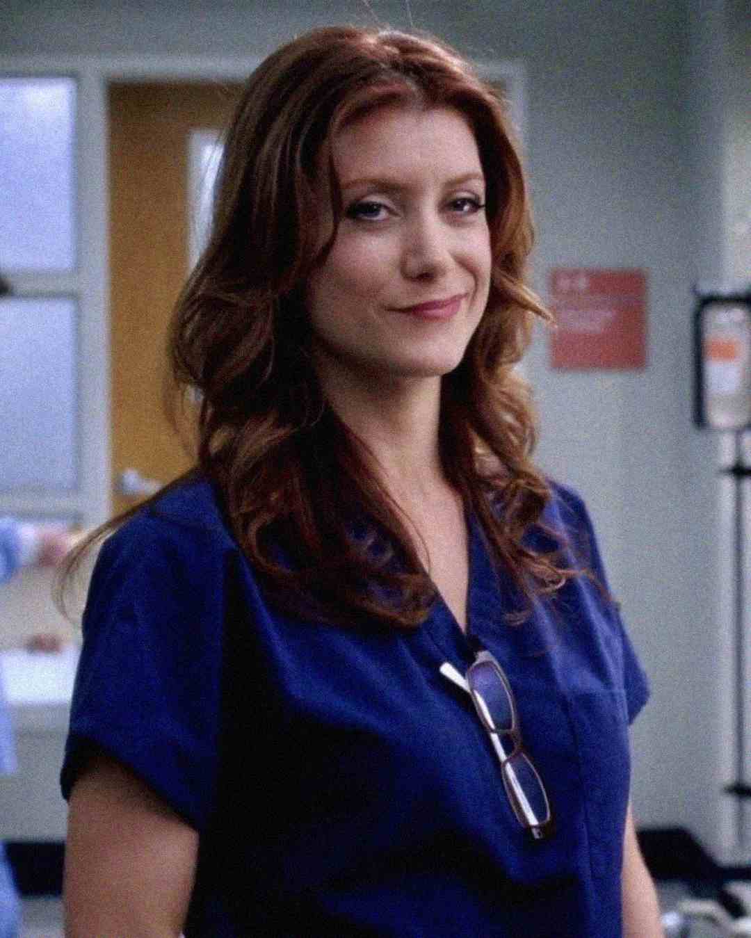 A woman with wavy brown hair, wearing blue medical scrubs and glasses hanging from the collar, stands in a hospital setting, smiling slightly.