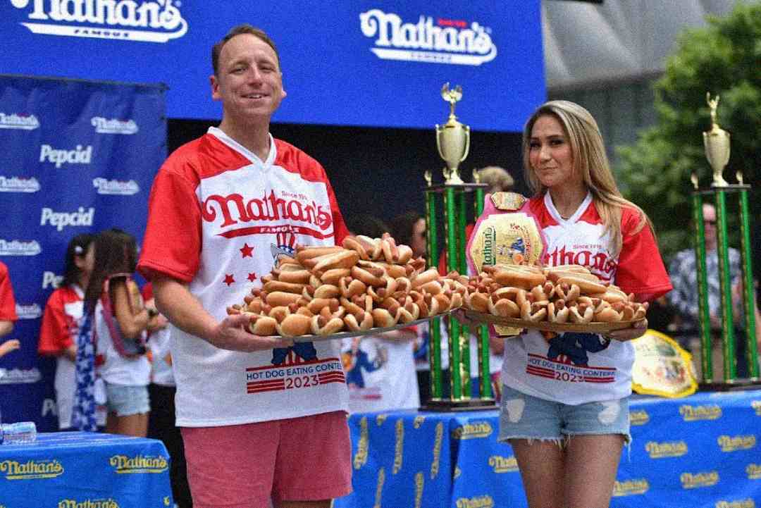 Two contestants at Nathan’s Famous Hot Dog Eating Contest hold large trays of hot dogs. They wear event jerseys and stand in front of trophies and a blue event backdrop. One contestant has a championship belt.