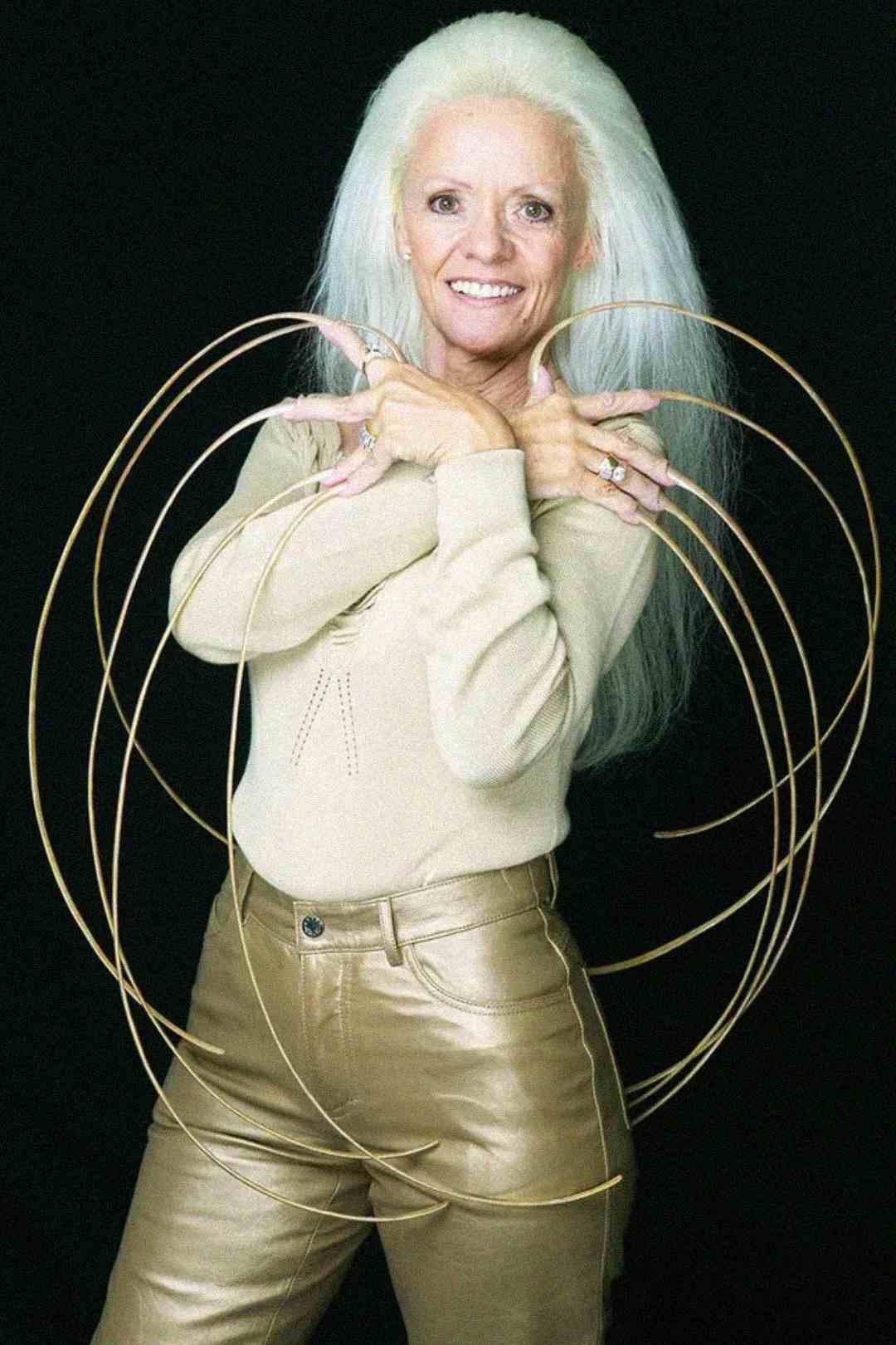 A smiling woman with long white hair poses in gold pants and a beige top, displaying extremely long, curved fingernails on both hands against a black background.
