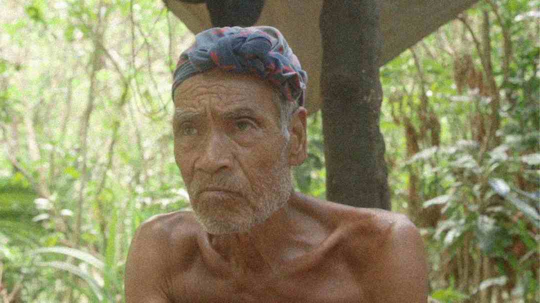 An elderly man with a gray beard and mustache sits shirtless outdoors, wearing a blue and red patterned headscarf. Lush green foliage and trees fill the background.