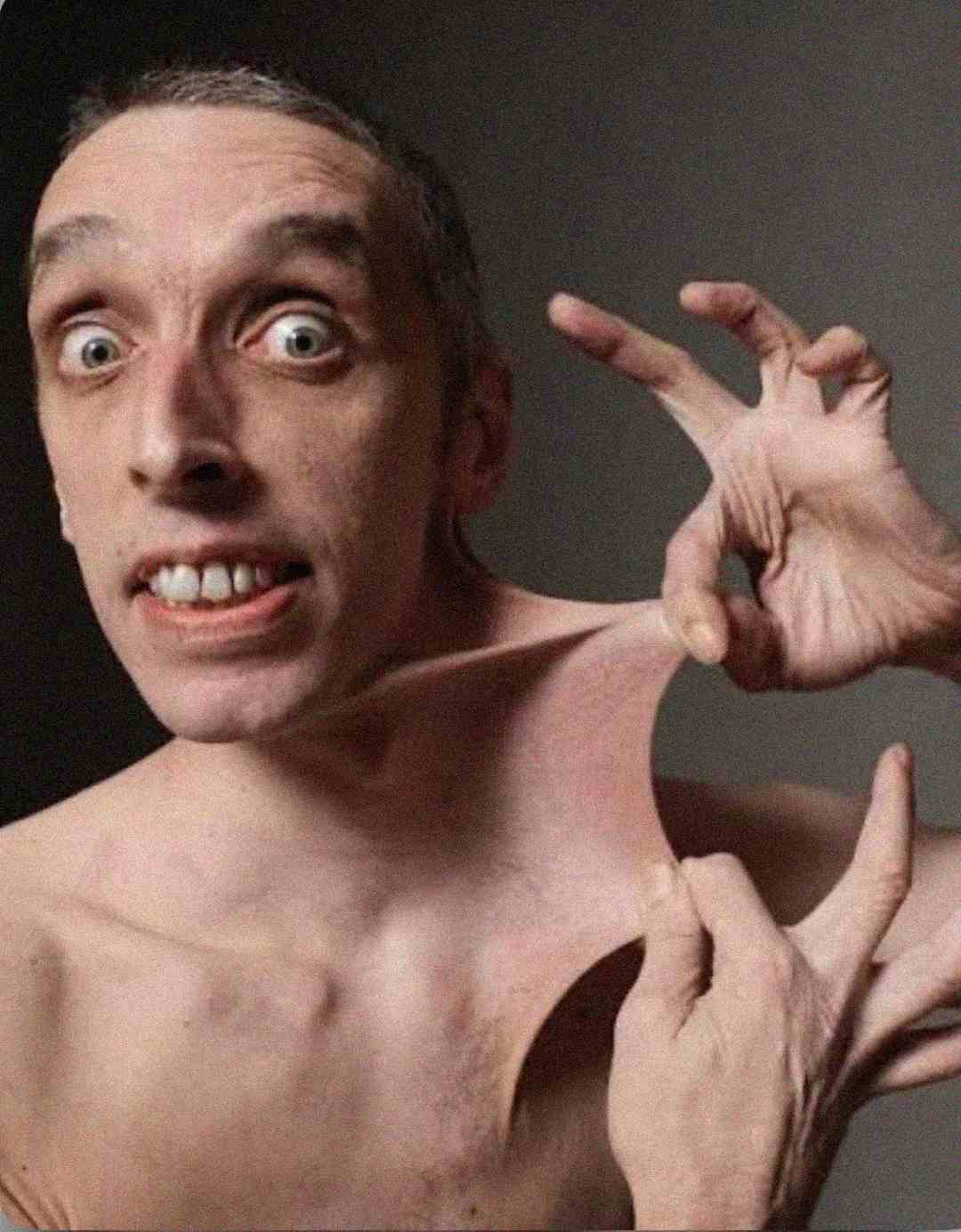 A shirtless person with wide eyes stretches the skin on their neck a long way using their hand, showcasing extremely flexible skin against a plain background.