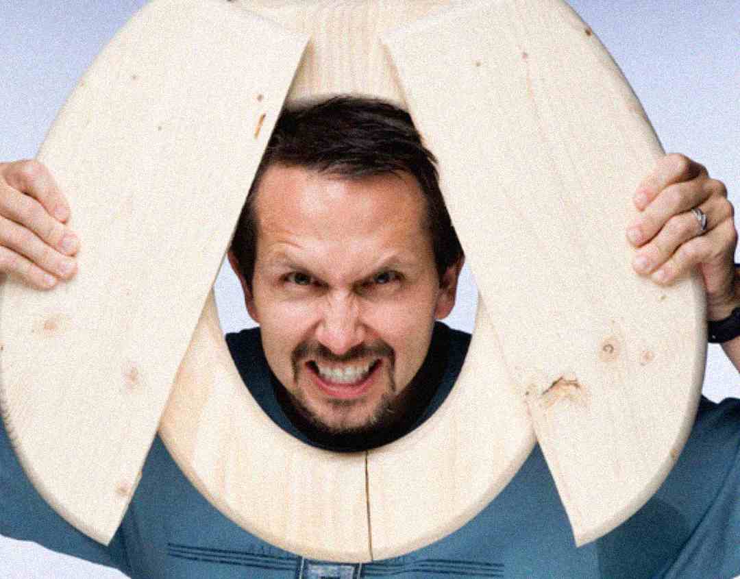 A man smiles with his teeth showing while holding a wooden toilet seat around his head, as if framing his face.