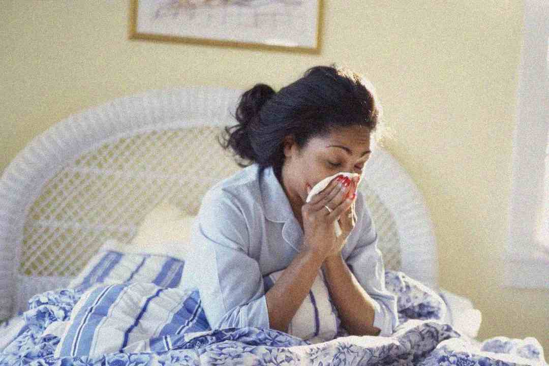 A woman sits up in bed wearing pajamas, holding a tissue to her nose as if sneezing or blowing her nose. The bedding is blue and white, and sunlight streams in through a nearby window.