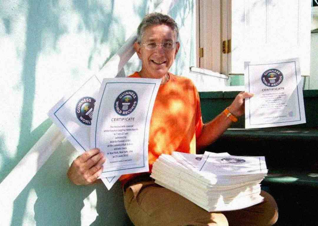A man in an orange shirt sits on outdoor steps, smiling and holding several Guinness World Records certificates, with a large stack of similar certificates on his lap. Sunlight and shadows fall across the scene.